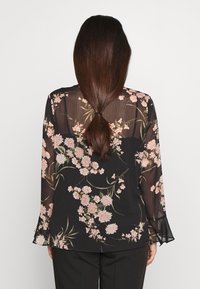 Svart genomskinlig blus med blommönster i rosa och grönt, långa ärmar med trumpetärmar. Slät textur, lätt design.