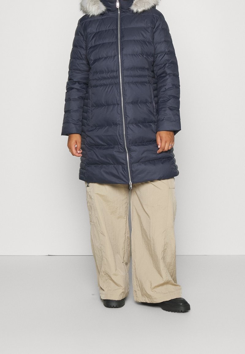 Marineblauer Steppmantel mit pelzbesetzter Kapuze, durchgehendem Reißverschluss und langen Ärmeln. Getragen über beigen, locker sitzenden Hosen und schwarzem Schuhwerk.