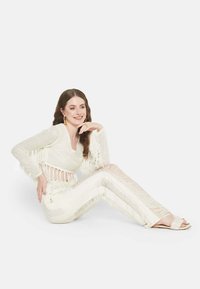 Weißes Fransenoberteil mit V-Ausschnitt, kombiniert mit Hosen mit Fransenakzenten und vertikalen Streifen. Das Model trägt flache Sandalen und auffällige Ohrringe.