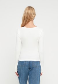 Γυναίκα με μακριά ξανθά μαλλιά που φορεί λευκή μπλούζα μακρυμάνικη και μπλε τζιν, στέκεται με την πλάτη προς το μέρος μας μπροστά σε ένα απλό φόντο.