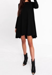 Abito lungo nero a maniche lunghe con una silhouette A-line morbida, che drappeggia fino a metà coscia. Abbinato a stivaletti neri alla caviglia con dettagli dorati sulla punta.