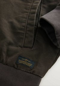 Gros plan sur la manche d'une veste marron foncé avec poignet côtelé et étiquette noire indiquant "Standard Issue Manufactured Goods Estd 1982."
