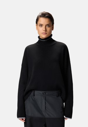 Maglione nero in maglia con colletto alto a coste, spalle scese e maniche ampie. Abbinato a pantaloni scuri con una texture liscia.