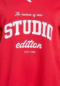 Červené bavlnené tričko s výrazným bielym textom: "STUDIO edícia," "Podstata prítomnosti," a "EST.1995." Výstrih do V s krátkymi rukávmi.