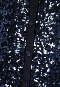 Tumš zilā krāsa, sequinam rotāta, aizpogājama apģērba gabals, kas noformēts ar apaļiem spīdumiem uz teksturēta auduma, ar četriem melniem pogām gar pogu rindu.