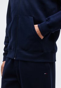 Άνδρας που φοράει σκούρα μπλε φούτερ με φερμουάρ και ταιριαστό παντελόνι, με το χέρι στην τσέπη του φούτερ, με μικρό κόκκινο, λευκό και μπλε λογότυπο στο παντελόνι.