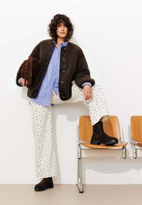 Veste brun texturé, chemise bleu clair à boutons, pantalon crème avec un motif floral bleu marine, et bottines noires en cuir. Cheveux bouclés.