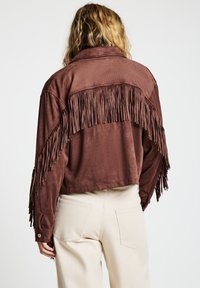 Braune Wildlederjacke mit Fransen über dem oberen Rücken. Verfügt über einen Kragen und geknöpfte Manschetten. Kombiniert mit hellen Hosen.