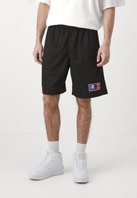 Bermudas deportivas de malla negras con una cinturilla elástica y bolsillos laterales, que presentan un parche con un logo colorido en la pierna izquierda.