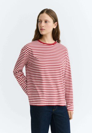 Jeune femme à la peau claire et aux longs cheveux bruns porte un t-shirt à manches longues rayé rouge et blanc et un jean bleu foncé, regardant vers sa gauche.