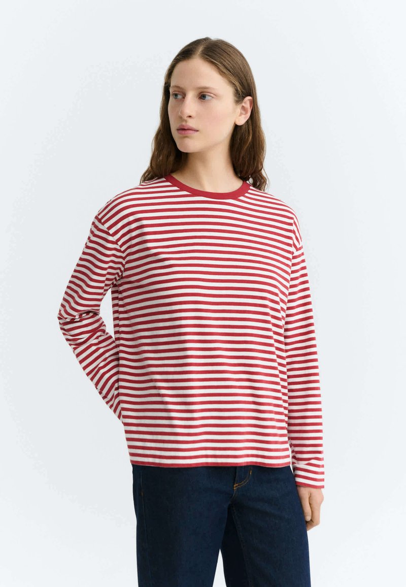 Jeune femme à la peau claire et aux longs cheveux bruns porte un t-shirt à manches longues rayé rouge et blanc et un jean bleu foncé, regardant vers sa gauche.