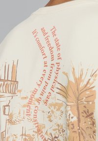 Crème katoenen T-shirt met een vervaagde print in perzik- en bruintinten, waarop abstracte ontwerpen en tekst in een gebogen indeling zijn weergegeven.