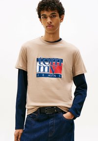 Ung mand iført en beige Tommy Jeans t-shirt over en navy langærmet trøje, med mørkeblå jeans og sort bælte, stående mod en hvid baggrund.