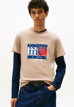 Junger Mann trägt ein beiges Tommy Jeans T-Shirt über einem navyfarbenen Langarmshirt, kombiniert mit dunkelblauen Jeans und einem schwarzen Gürtel, während er vor einem weißen Hintergrund steht.