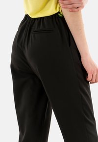 Pantaloni neri con una texture liscia, vita elasticizzata e due tasche posteriori decorative; abbinati a una maglia gialla brillante.