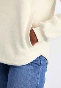 Pullover in pile color crema con una texture morbida, dotato di polsini a costine e una tasca laterale. Abbinato a jeans in denim blu.