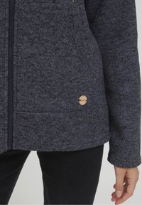 Giacca effetto tessuto blu scuro con zip, dotata di un logo in legno e due tasche laterali. Tessuto morbido con una vestibilità casual.