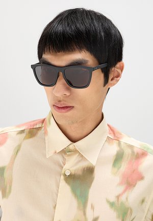 Giovane uomo con capelli neri lisci che indossa occhiali da sole rettangolari neri e una camicia chiara con bottoni con motivi ad acquerello verdi e rosa.