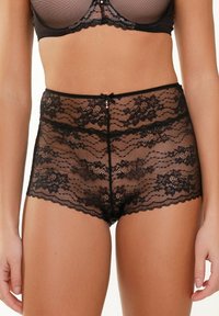 Culotte taille haute en dentelle noire avec des motifs floraux, des bords festonnés, un accent en nœud central et une texture transparente.