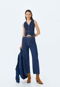Chaleco de mezclilla con escote en V y cierre de cinco botones, combinado con pantalones de mezclilla de pierna ancha. La modelo sostiene una chaqueta a juego, llevando botas de tacón marrones.