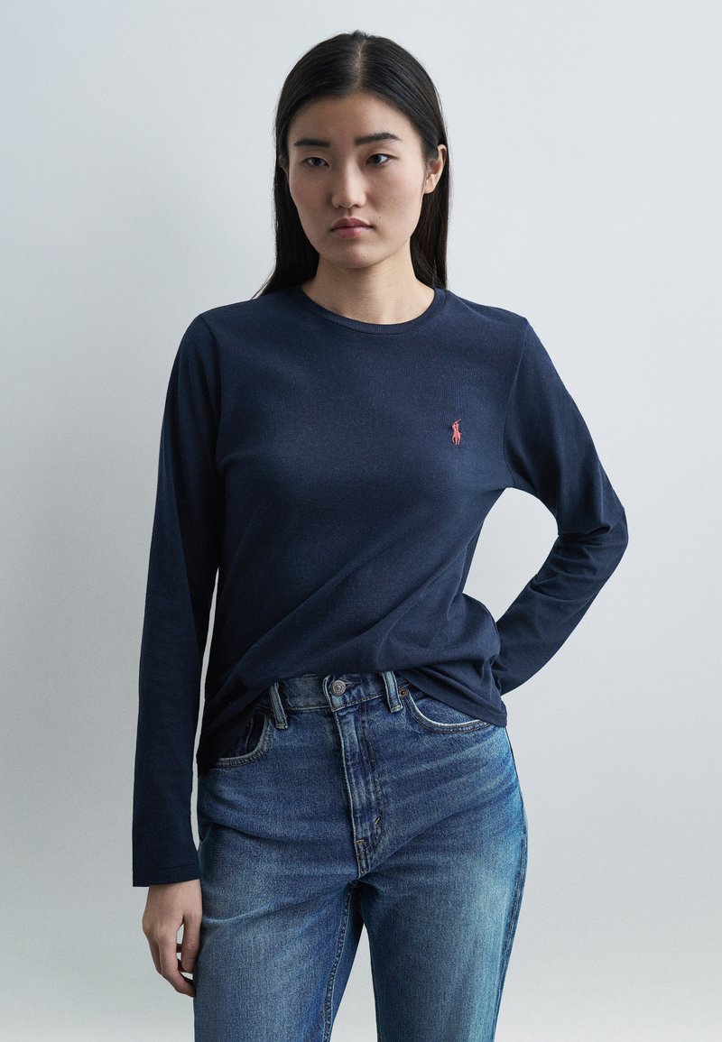 Γυναίκα με μακριά σκούρα μαλλιά, φοράει μπλε σκο ύρο πουκάμισο με μακριά μανίκια και ένα μικρό κόκκινο λογότυπο και μπλε τζιν, στέκεται μπροστά σε απλό γκρι φόντο.