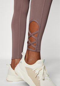 Legginsy sportowe w stonowanym mauve z bocznym wycięciem i krzyżującymi się detalami, w połączeniu z białymi trampkami z beżowymi akcentami.