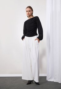 Zwarte blouse met lange mouwen en sweetheart-halslijn, gecombineerd met hoge witte broek met wijde pijpen en zwarte stiletto-hakken. Effen achtergrond.