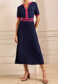 Robe marine à manches courtes, avec un col et une ceinture en scallop rose, dotée d'une jupe plissée. Des sandales à talons bruns complètent le look.