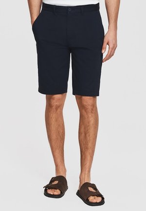 Mężczyzna noszący ciemnogranatowe spodenki do kolan z przednimi kieszeniami i ciemnobrązowe sandały na klamrę, stojący na jednolitym tle.