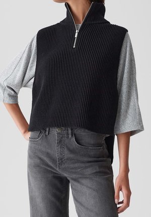 Femme portant une veste sans manches noire côtelée avec fermeture éclair sur une chemise grise à manches trois-quarts et un jean gris, debout avec une main sur la hanche.