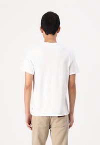 Tricou alb din bumbac cu mâneci scurte, decolteu rotund și un design simplu, asortat cu pantaloni bej ce au detalii de cusătură la buzunarul din spate.