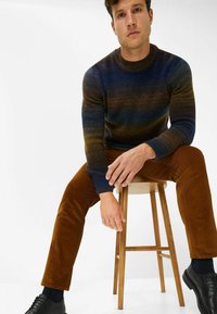 Strickpullover mit blau-braunem Farbverlauf, runder Ausschnitt; kombiniert mit braunen Cordhosen und schwarzen Schuhen, sitzt auf einem Holzstuhl.