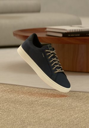 Marineblå sneaker med hvid sål og grå snørebånd, svævende over et beige tekstureret tæppe, med et rundt bord i træ og bøger i baggrunden.