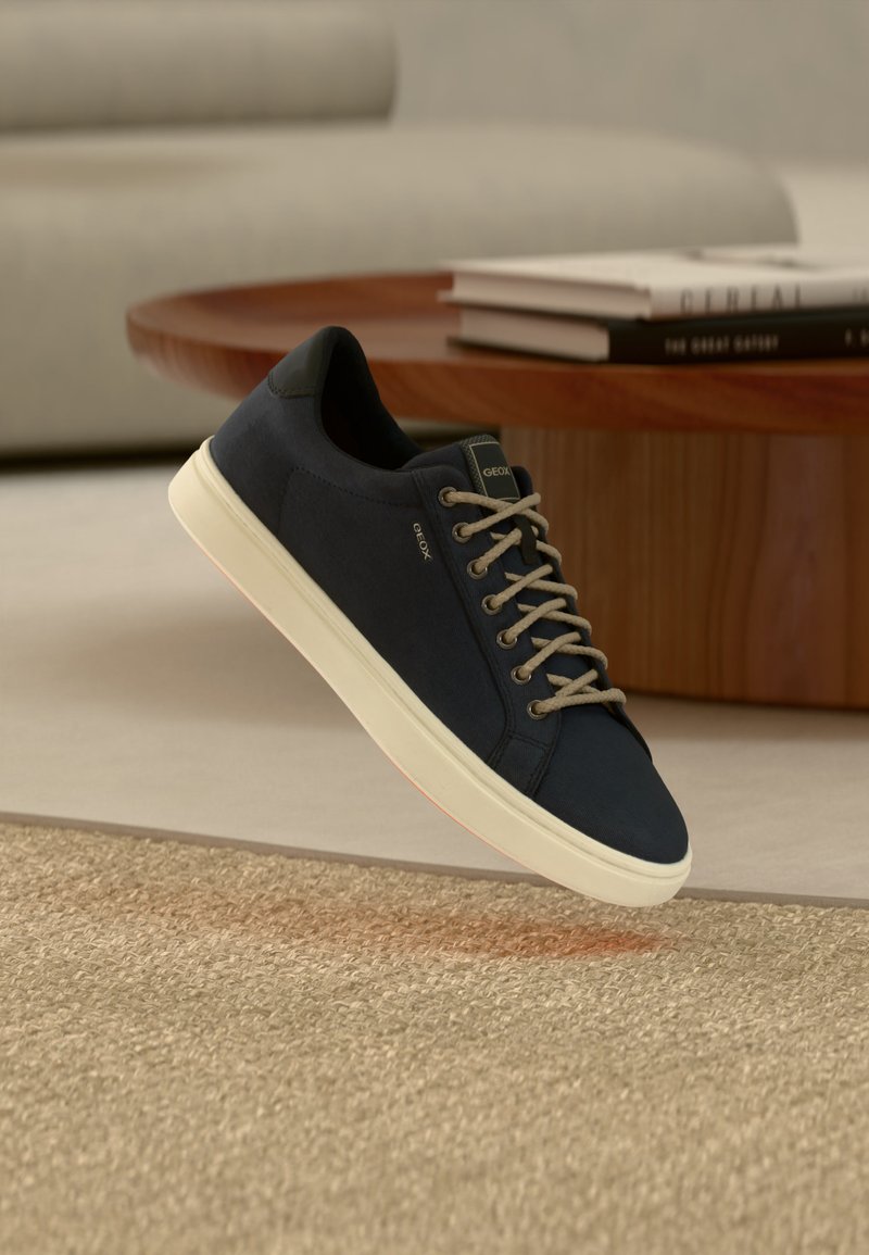 Σκούρο μπλε αθλητικό παπούτσι με λευκή σόλα και γκρι κορδόνια, κρεμασμένο πάνω από μια μπεζ υφασμάτινη μοκέτα, με στρογγυλό ξύλινο τραπέζι και βιβλία στο φόντο.