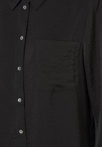 Chemise noire à manches longues avec un tissu texturé, une poche poitrine et des boutons gris. Le col est standard. Le design global est simple et classique.