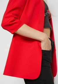 Röd kavaj i ett texturerat tyg, avslappnad passform och trekvartslånga ärmar. Har en sidoficka och släta kanter, matchad med svarta jeans.