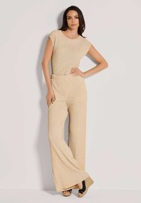 Beige gebreide jumpsuit met korte mouwen, broekspijpen met wijd uitlopende pijpen, gestructureerde stof en een aangesloten taille. Gecombineerd met zwarte sleehakken.