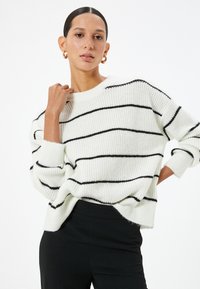 Pull en tricot blanc avec des rayures horizontales noires, doté d'un col rond et d'une coupe oversized, porté avec un pantalon noir. Tissu texturé.