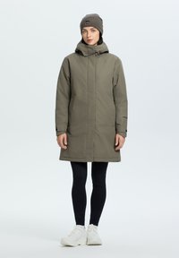 Oliivinvihreä pitkä parkatakki hupulla, jossa on vetoketju ja napit, yhdistetty mustiin legginsseihin ja valkoisiin lenkkareihin. Päällä neulottu pipo.