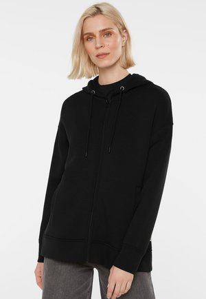Blond woman wearing a black zip-up hoodie with drawstrings and gray pants, standing against a plain white background.