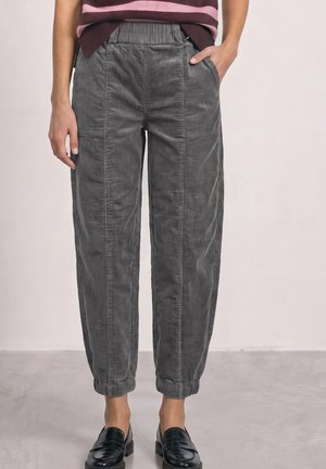 Femme portant un pantalon en velours côtelé gris avec des poignets élastiques et des mocassins noirs, debout avec une main dans la poche.
