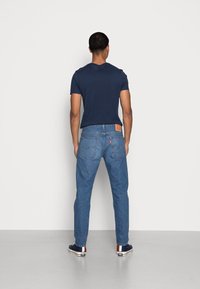Camiseta azul oscura combinada con jeans de mezclilla azul claro. Los jeans tienen un parche de cuero marrón y múltiples bolsillos. Calzado casual completa el look.