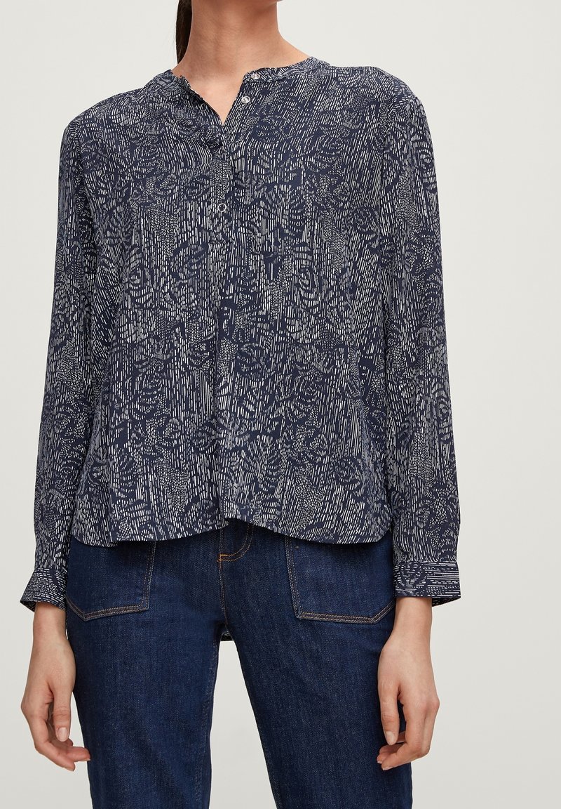 Femme portant un chemisier à manches longues à motifs bleu marine et blanc associé à un jean denim bleu foncé, debout sur un fond clair.