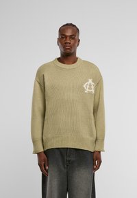 Olivengrüner Strickpullover mit geripptem Halsausschnitt und Bündchen, mit einem weißen Logo-Detail auf der Brust, kombiniert mit ausgewaschenen grauen Hosen.