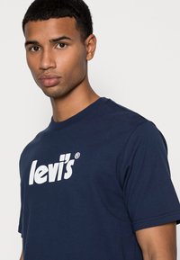 Navy sininen puuvillainen t-paita lyhyillä hihoilla, jossa on suuri valkoinen "Levi's" -logo. Pyöreä pääntie, sileä rakenne.