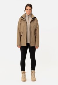 Giacca impermeabile color cammello con zip frontale, dettagli neri e molteplici tasche, indossata sopra un fleece grigio chiaro, abbinata a leggings neri e stivali beige.