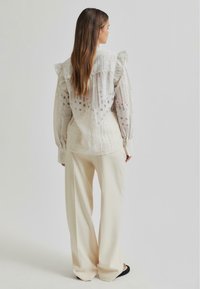 Lightweight, cream blouse with textured fabric, featuring sheer dotted patterns and ruffled shoulders, paired with flowy, beige trousers.