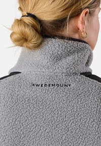 Fleecejacka i ljusgrått med en texturerad yta. Har en hög krage och svart kant, med "SWEDEMOUNT" logotyp broderad på ryggen.