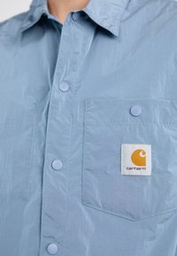 Chemise bleu clair à manches longues en tissu lisse et brillant. Présente une poche poitrine et un patch logo Carhartt orange et blanc bien visible.