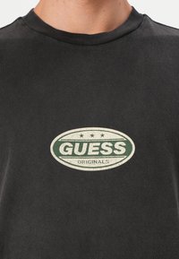 Svart bomulls t-skjorte med en grønn oval logo med teksten "GUESS ORIGINALS," samt en teksturert hvit kant og tre stjernesymboler over.